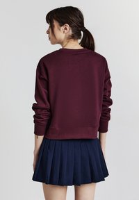 Donna con capelli scuri raccolti in una coda di cavallo, che indossa un maglione a maniche lunghe color bordeaux e una gonna plissettata blu navy, rivolta di spalle rispetto alla fotocamera.