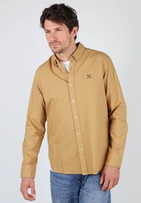 Chemise à manches longues, boutons sur le devant, de couleur marron clair avec un col pointu, boutons blancs et un petit motif brodé sur la poitrine. Coupe décontractée.