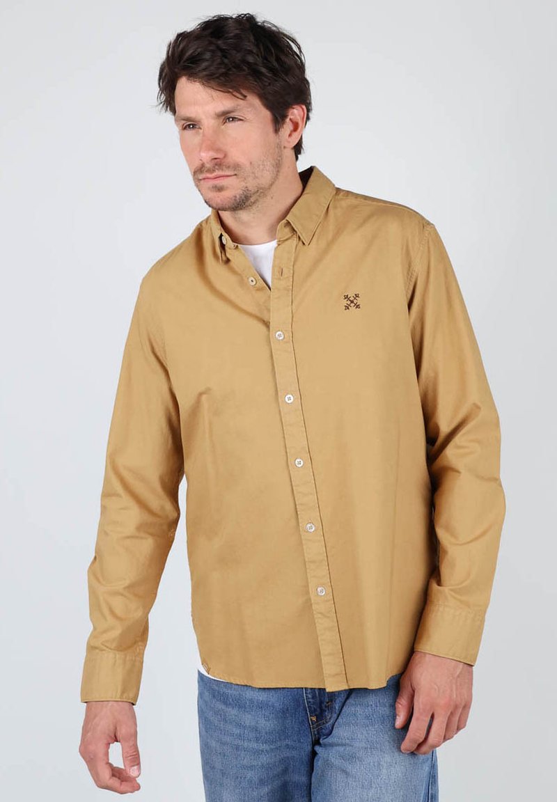 Chemise à manches longues, boutons sur le devant, de couleur marron clair avec un col pointu, boutons blancs et un petit motif brodé sur la poitrine. Coupe décontractée.