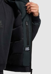 Svart jakkeinnside med polstret isolasjon, glidelåslomme og tekstillapp. Yttermaterialet ser glatt ut, med ribbestrikkede mansjetter.