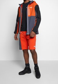 Functionele outdooroutfit met een waterdichte jas in oranje en grijs, gecombineerd met oranje shorts en zwarte wandelschoenen.