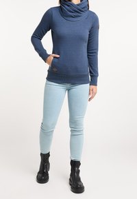 Námořnická modrá mikina s vysokým límcem, dlouhými rukávy a žebrovanými manžetami, obsahující kapsu a hnědou nášivku s logem. Spárována se světlými modrými džíny.