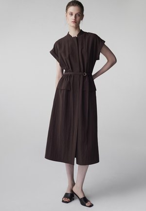 Mujer de pie con la mano en la cadera, vestida con un vestido midi marrón con cinturón, mangas cortas y mules negros de tacón abiertos en los dedos.