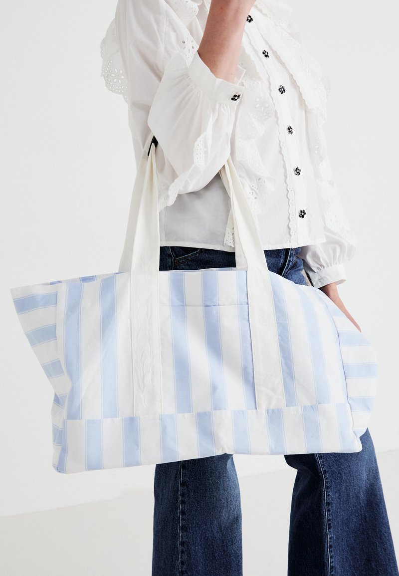 Pessoa vestindo uma blusa branca e jeans azuis segura uma grande bolsa de lona listrada em azul claro e branco, com alças longas.