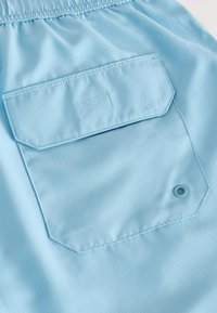 Tissu bleu clair à texture lisse comportant une poche rectangulaire plaquée et un œillet circulaire. Taille détendue avec élastique.