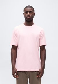 Άνδρας που φοράει ανοιχτό ροζ κοντομάνικη μπλούζα και μπεζ παντελόνι, στέκεται μπροστά σε απλή ανοιχτογκρι φόντο.