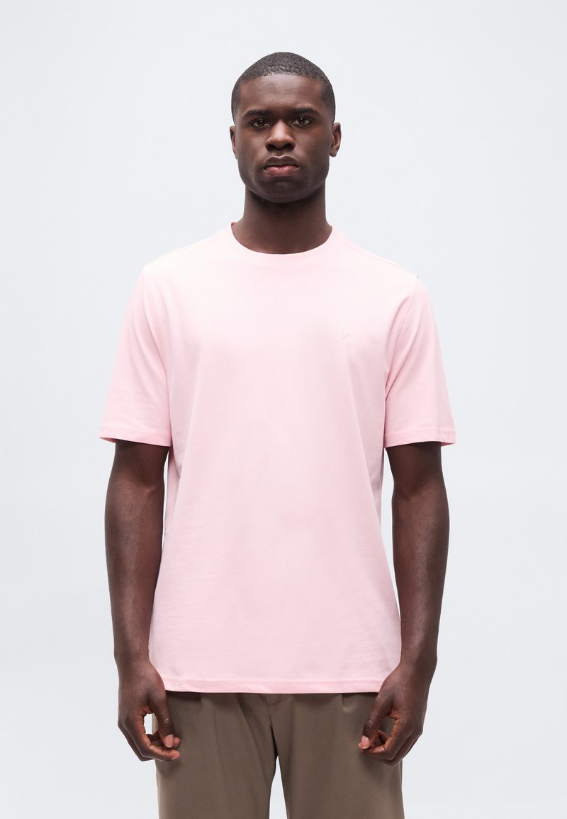 Άνδρας που φοράει ανοιχτό ροζ κοντομάνικη μπλούζα και μπεζ παντελόνι, στέκεται μπροστά σε απλή ανοιχτογκρι φόντο.