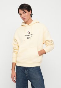 Sudadera amarilla de algodón con un bolsillo delantero, que presenta el texto "tommy girl" y un logo de estrella en negro y rojo. Fit casual, puños acanalados.