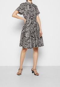 Frau in einem knielangen Kleid mit schwarz-weißem Muster, kurzen Ärmeln und schwarzen Riemchen-Absatzsandalen, die mit einer Hand auf der Hüfte posiert.