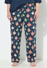 Pantalones de pijama de color azul marino con patrones de adornos navideños en rojo, verde y blanco, usados por una persona descalza de pie contra un fondo blanco.