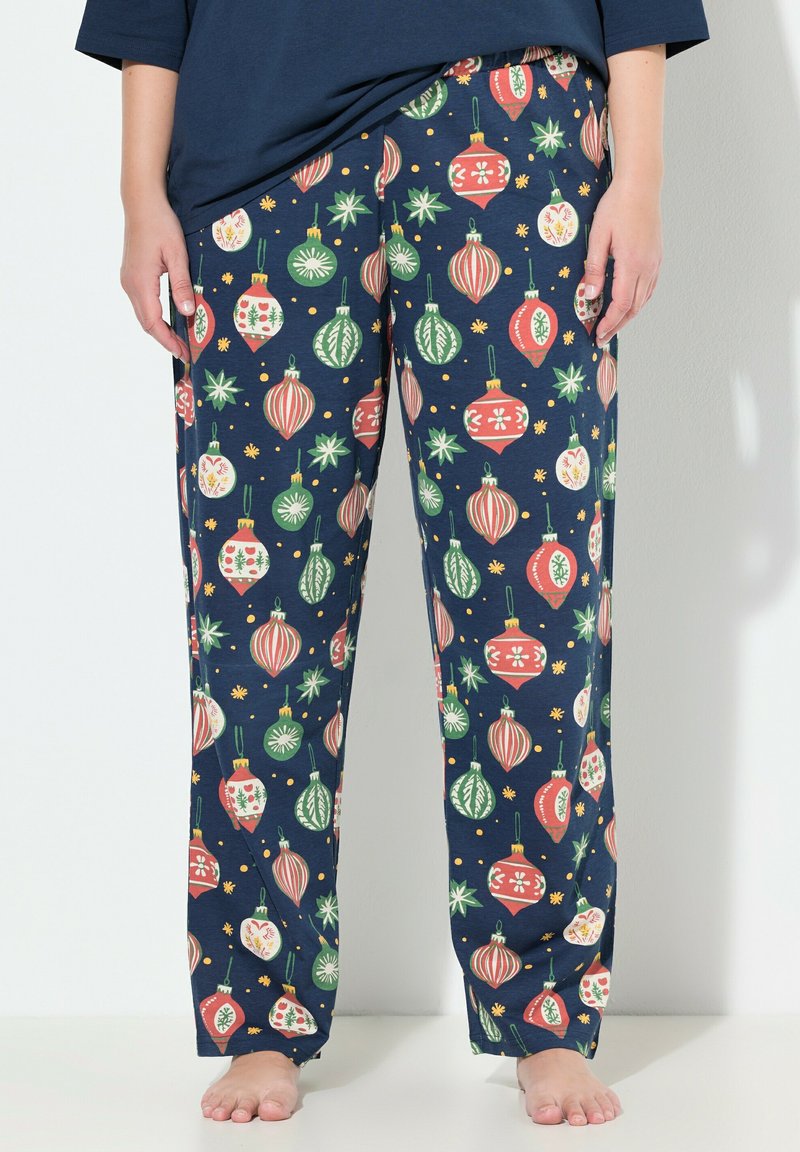 Pantalones de pijama de color azul marino con patrones de adornos navideños en rojo, verde y blanco, usados por una persona descalza de pie contra un fondo blanco.