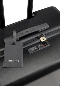 Zwarte harde koffer met een gestructureerde oppervlakte, combinatielock, gouden accenten en een zwarte bagagetag met de tekst "ETERNITIVE" ingeperst.