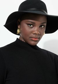 Mustat neulepaidat, joissa on korkea kaula-aukko, yhdistettynä leveäreunaiseen mustaan huopahatun. Koristeena tekstuuriset kultakorvakorut, joissa on veistoksellinen muotoilu.