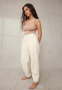 Vaalean beige hihaton paita yhdistettynä valkoisiin collegehousuihin, joissa on joustava vyötärönauha ja hihnat. Collegehousuissa on pieni "self." logo.