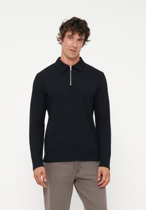 Homme aux cheveux bouclés portant un shirt noir à manches longues avec une fermeture éclair à l'avant et un pantalon gris, debout devant un fond blanc uni.