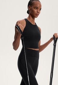 Crop top atletico nero con tessuto liscio e design con schiena a corsetto. Leggings neri con cuciture testurizzate. Individuo tiene delle bande di resistenza.