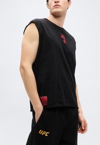 Μαύρο αμάνικο μπλουζάκι με κόκκινο λογότυπο, κατασκευασμένο από βαμβάκι. Συνδυασμένο με μαύρο παντελόνι που έχει χρυσό λογότυπο "UFC" και πλαϊνές τσέπες.