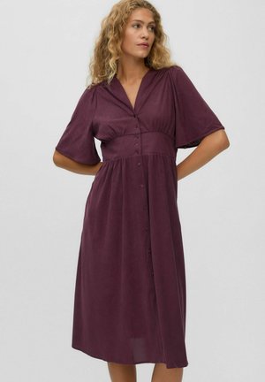 Woman with curly hair wearing a buttoned, deep purple midi dress with short wide sleeves and hands resting on her hips.