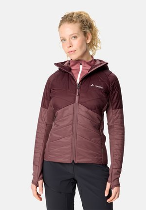 Frau mit lockigem blondem Haar, die eine zweifarbige Mauve-Bordeaux Kapuzenjacke und schwarze Hose trägt, steht vor einem schlichten hellen Hintergrund.