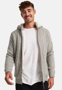 Cárdigan de punto gris claro con capucha, que presenta un patrón de punto trenzado, cierre de cremallera y puños acanalados. Se lleva encima de una camiseta blanca.