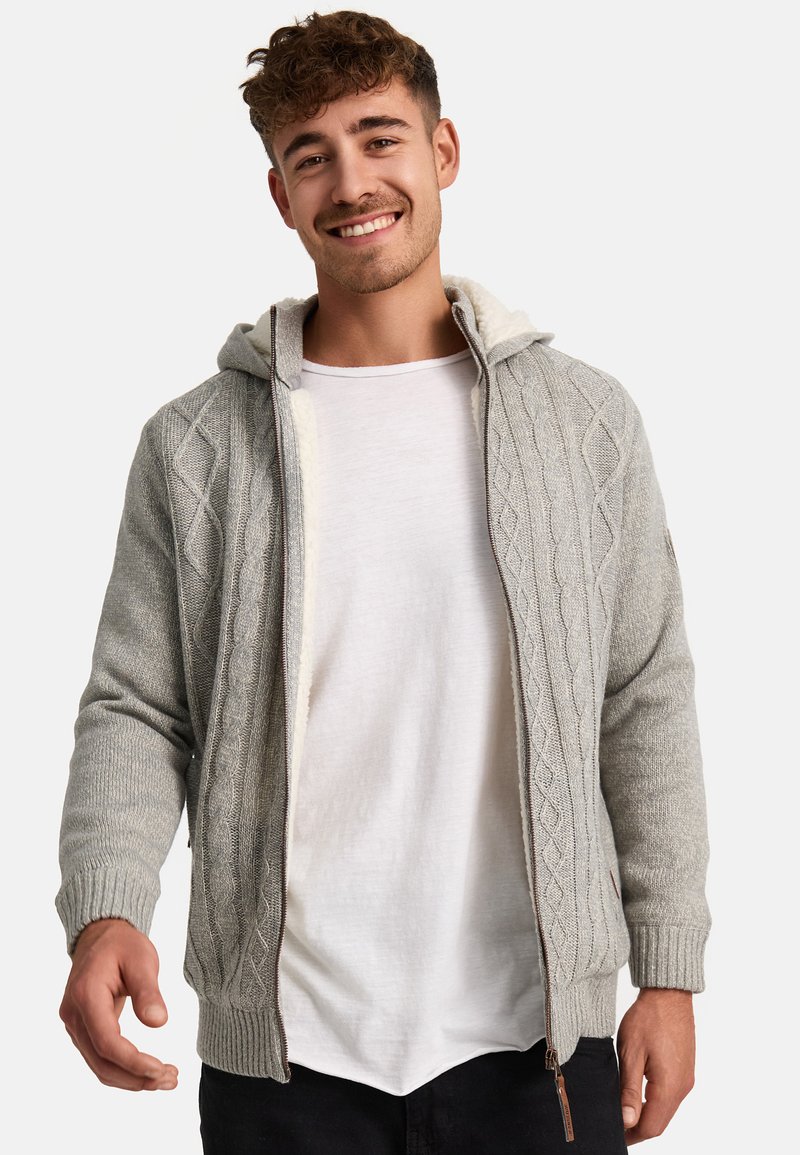Cárdigan de punto gris claro con capucha, que presenta un patrón de punto trenzado, cierre de cremallera y puños acanalados. Se lleva encima de una camiseta blanca.
