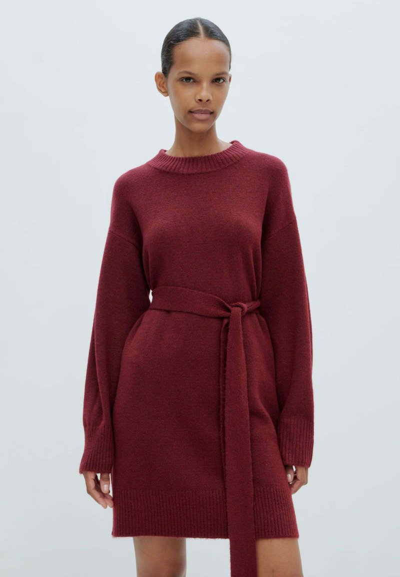 Strickkleid in Dunkelrot, mit hohem Ausschnitt, langen Ärmeln und einem passenden Gürtel, der in der Taille gebunden ist. Rippstruktur an den Ärmeln und am Saum.