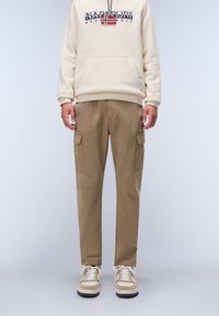 Beige hættetrøje med frontlomme og logo, parret med lysebrune cargo-bukser med sidelommer, og beige/creme sneakers.