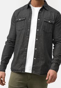 Camisa de color gris oscuro hecha de tela de mezclilla, con un frente abotonado, dos bolsillos en el pecho y una textura desgastada con detalles de botones a presión.
