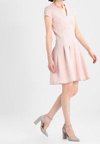 Helles rosa Kleid mit kurzen Ärmeln, V-Ausschnitt, strukturiertem Muster, tailliertem Oberteil und weit ausgestellt Rock. Kombiniert mit grauen High Heels.