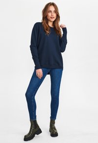 Tengerészak kék pulóver, hosszú ujjakkal, bordás mandzsettával és szegéllyel. Karcsú kék farmernadrággal és vastag, olívazöld bokacipővel párosítva.