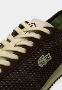 Καφέ αθλητικό παπούτσι με δίχτυ, ανοιχτοκίτρινες κορδόνια, πράσινη εσωτερική επένδυση και λογότυπο κροκόδειλου σε πράσινο χρώμα. Διαθέτει υφή στην επιφάνεια και μαξιλαρωμένο σχεδιασμό.