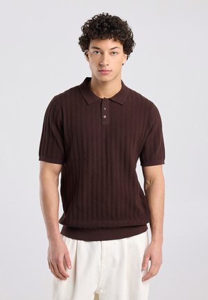 Jonge man met krullend haar, gekleed in een donkerbruin poloshirt met korte mouwen en een gestructureerd oppervlak, en losse witte broek, tegen een effen achtergrond.