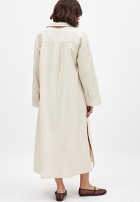 Pitkä, beige paitamekko kevyestä kankaasta, rento malli, takana laskos, sivuventtiilit ja nappikahvat. Yhdistetty tummanruskeisiin kenkiin.