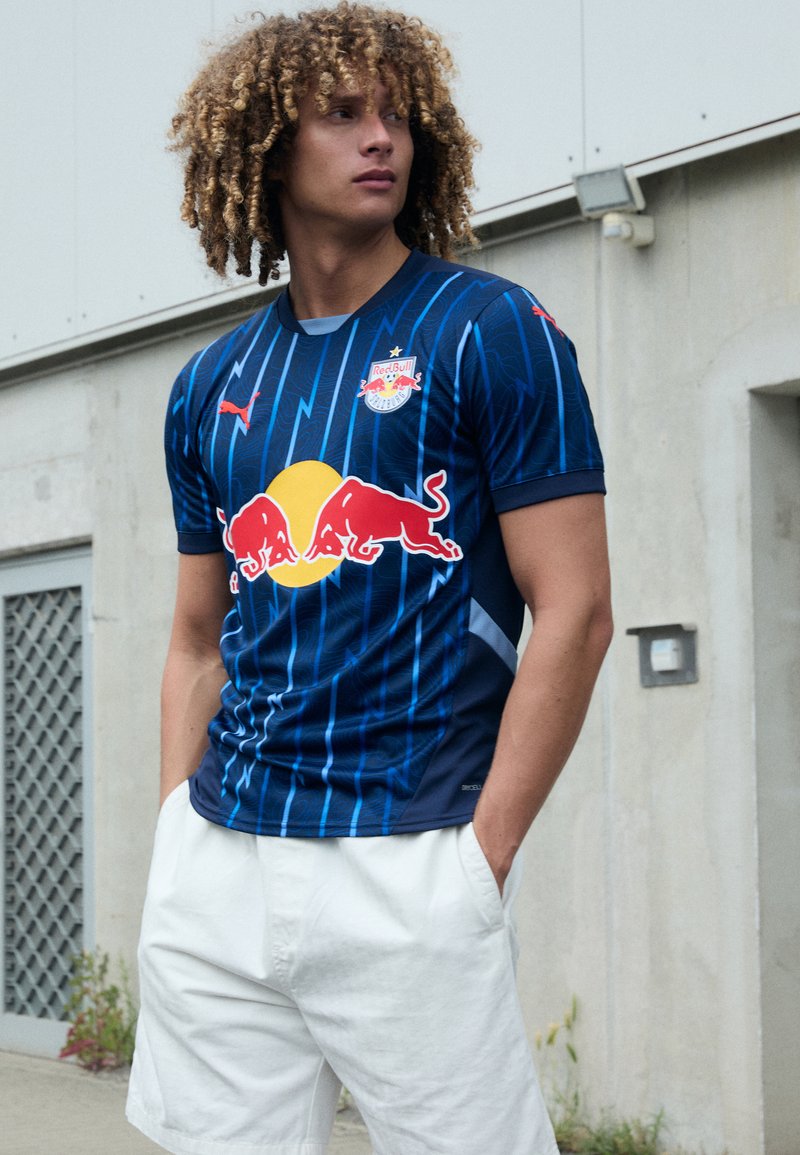 Maglia sportiva a righe blu con logo Red Bull, maniche corte, realizzata in materiale traspirante, abbinata a pantaloni bianchi. Vestibilità casual e atletica.