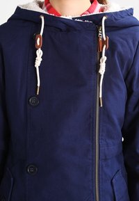 Giacca blu navy con cappuccio, chiusura a zip e due bottoni. Presenta lacci bianchi con dettagli marroni e una morbida fodera interna.