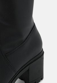 Černá kožená kotníková bota s texturovaným povrchem, robustním podpatkem a viditelnými švy na stranách. Silná, vzorovaná podešev.