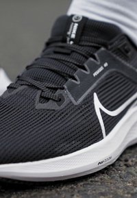 Svart idrettssko med teksturert overdel, hvit Nike swoosh og grå detaljer. Har polstret krage og synlig Air Zoom demping.