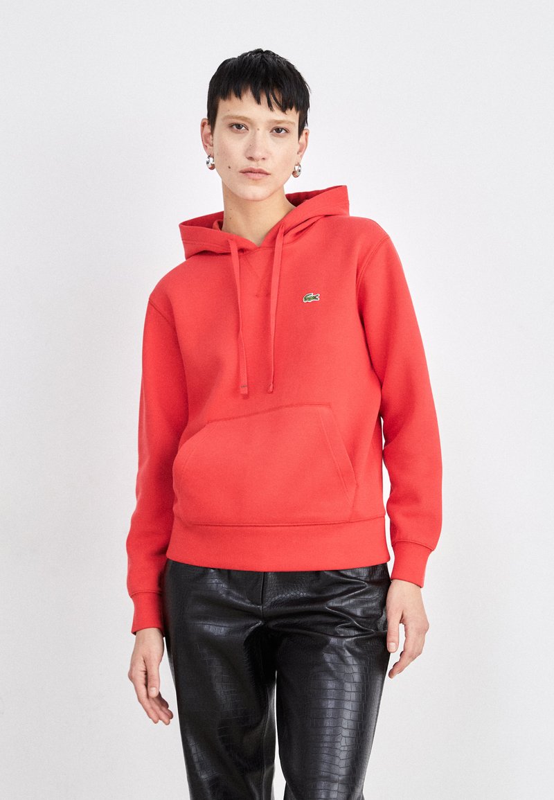Felpa con cappuccio rossa realizzata in materiale morbido, con tasca frontale e un piccolo logo verde. Abbinata a pantaloni neri lucidi e testurizzati.