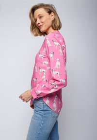 Langarmshirt aus pinkem Stoff mit einem Hundemuster. Verfügt über einen abgerundeten Saum und knöpfbare Manschetten. Kombiniert mit hellblauen Denim-Jeans.