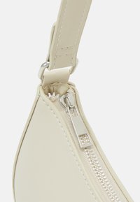 Světle béžový roh kožené tašky se stříbrným zipem, s hladkou texturou a poutkem pro popruh. Viditelný je jednoduchý design a čisté linie.