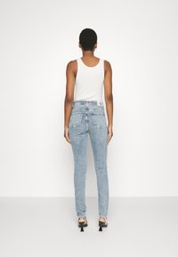 Jeans de mezclilla de tiro alto, de un azul claro, con una silueta ajustada, bolsillos traseros y una etiqueta de logo en la cintura. Se llevan con una camiseta sin mangas blanca.