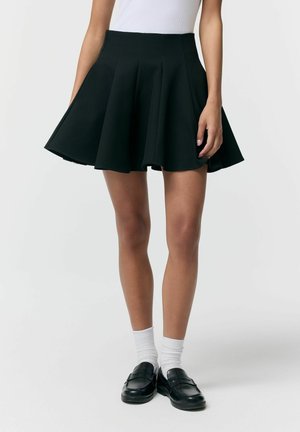 Kobieta ubrana w czarną plisowaną mini spódniczkę, białe skarpetki, czarne mokasyny i biały, bezrękawnik, stojąca na tle jednolitego tła.