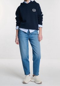 Sudadera con capucha azul marino con bolsillo frontal, logotipo blanco, superpuesta sobre una camisa de rayas azul claro. Jeans azul claro y zapatillas crema completan el conjunto.