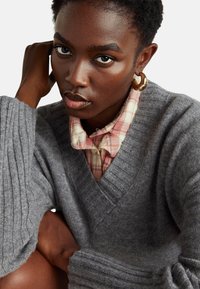 Šedý pletený svetr s ribbovaným designem a širokými rukávy, vrstvený přes růžovou kostkovanou košili, zdobený velkým kulatým náušnicí.