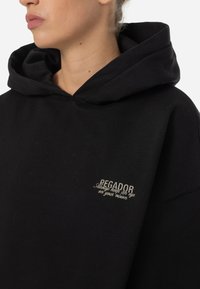 Felpa nera oversize in cotone, caratterizzata da un ampio cappuccio e una patch ricamata sulla parte anteriore con la scritta "PEGA dor" e una frase in colore chiaro.