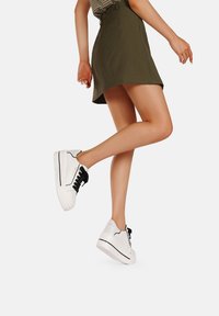 Sneakers bianchi con correttori neri, caratterizzati da una tomaia liscia, punta rotonda e suola spessa. L'outfit include una gonna verde e una maglietta a righe.