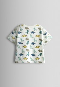 Biała, dziecięca koszulka z krótkim rękawem, ozdobiona kolorowymi, kreskówkowymi wzorami dinozaurów w kolorach niebieskim, żółtym, zielonym i brązowym na jednolitym tle.