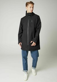 Schwarze wasserdichte Jacke mit einem Frontreißverschluss, verstellbare Kapuze und einer Brusttasche. Kombiniert mit hellblauen Jeans und weißen Sneakers.