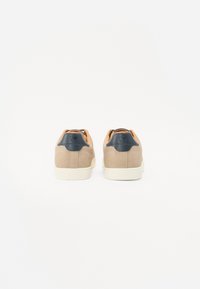 Beige sneakers with a dark blue heel accent, white rubber sole, and textured suede upper. Features contrasting tan interior and stitching.