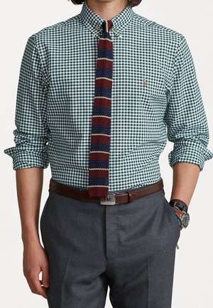 Man draagt een groen-wit geruit overhemd met opgerolde mouwen, gestreepte gebreide stropdas, bruine riem, donkergrijze broek en polshorloge met armbanden.